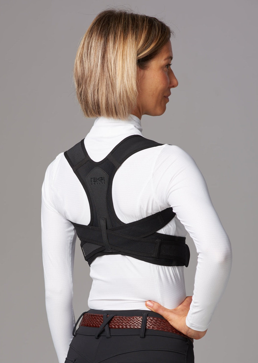 Female wearing dark riding pants, white tip and brown leather belt. She wears a black posture brace over the top of her white shirt.