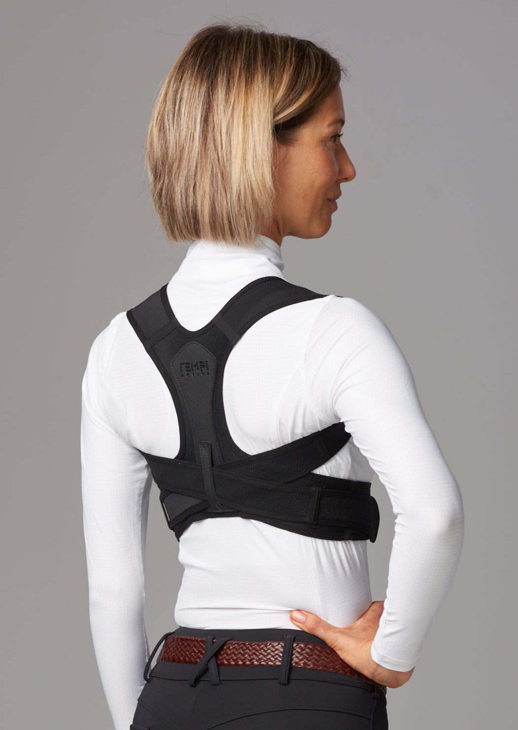 Female wearing dark riding pants, white top and brown leather belt and black posture brace facing side-ways.