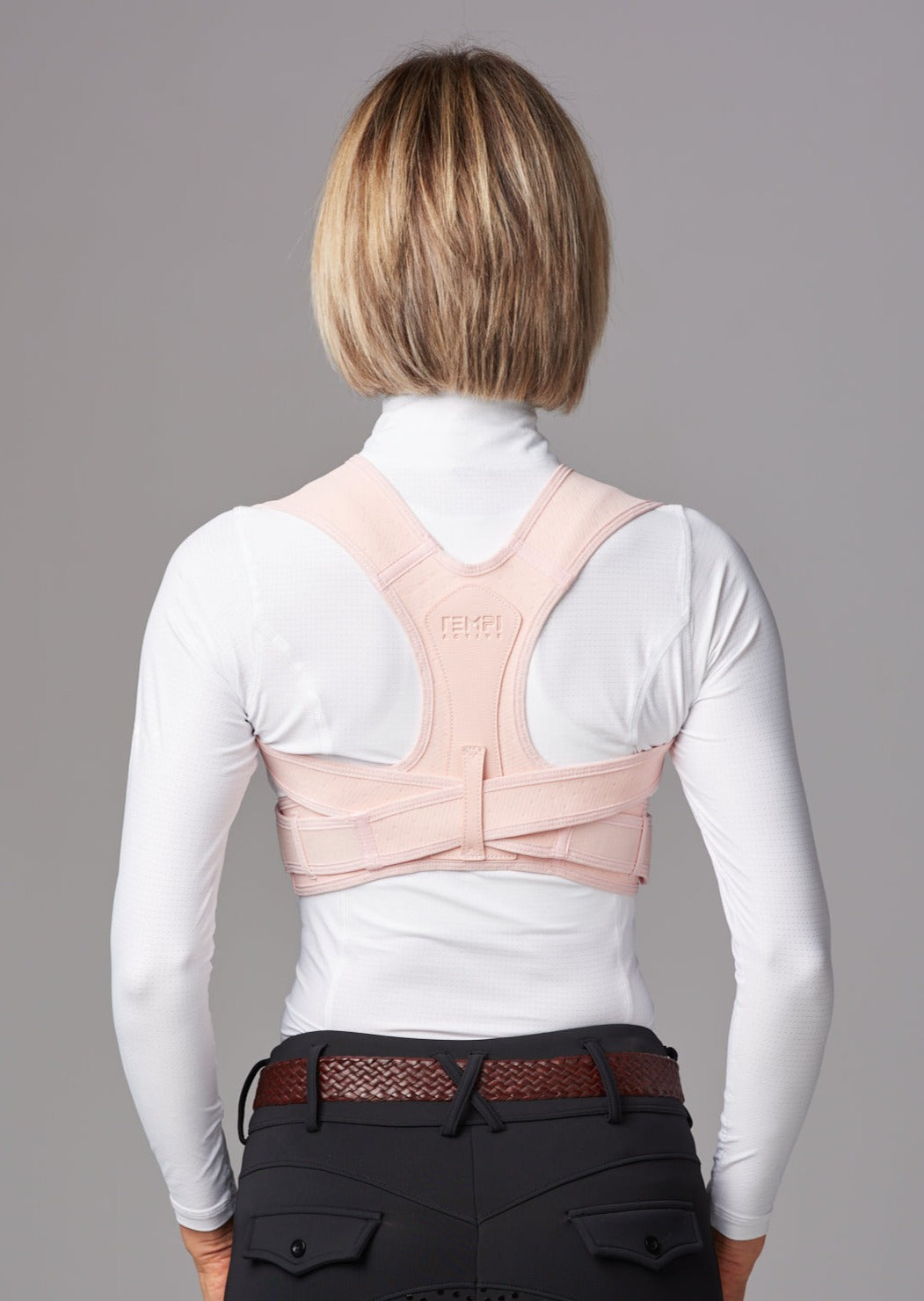 Female wearing dark riding pants, white tip and brown leather belt. She wears a pink posture brace over the top of her white shirt. She is facing away from the camera.