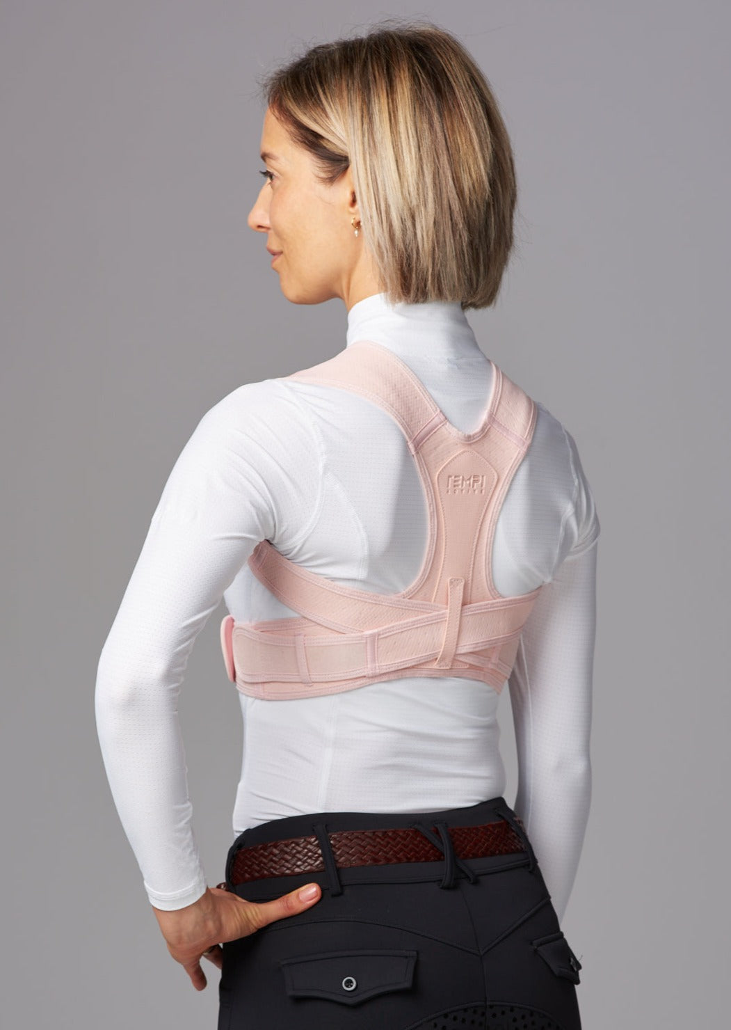 Female wearing dark riding pants, white tip and brown leather belt. She wears a pink posture brace over the top of her white shirt.