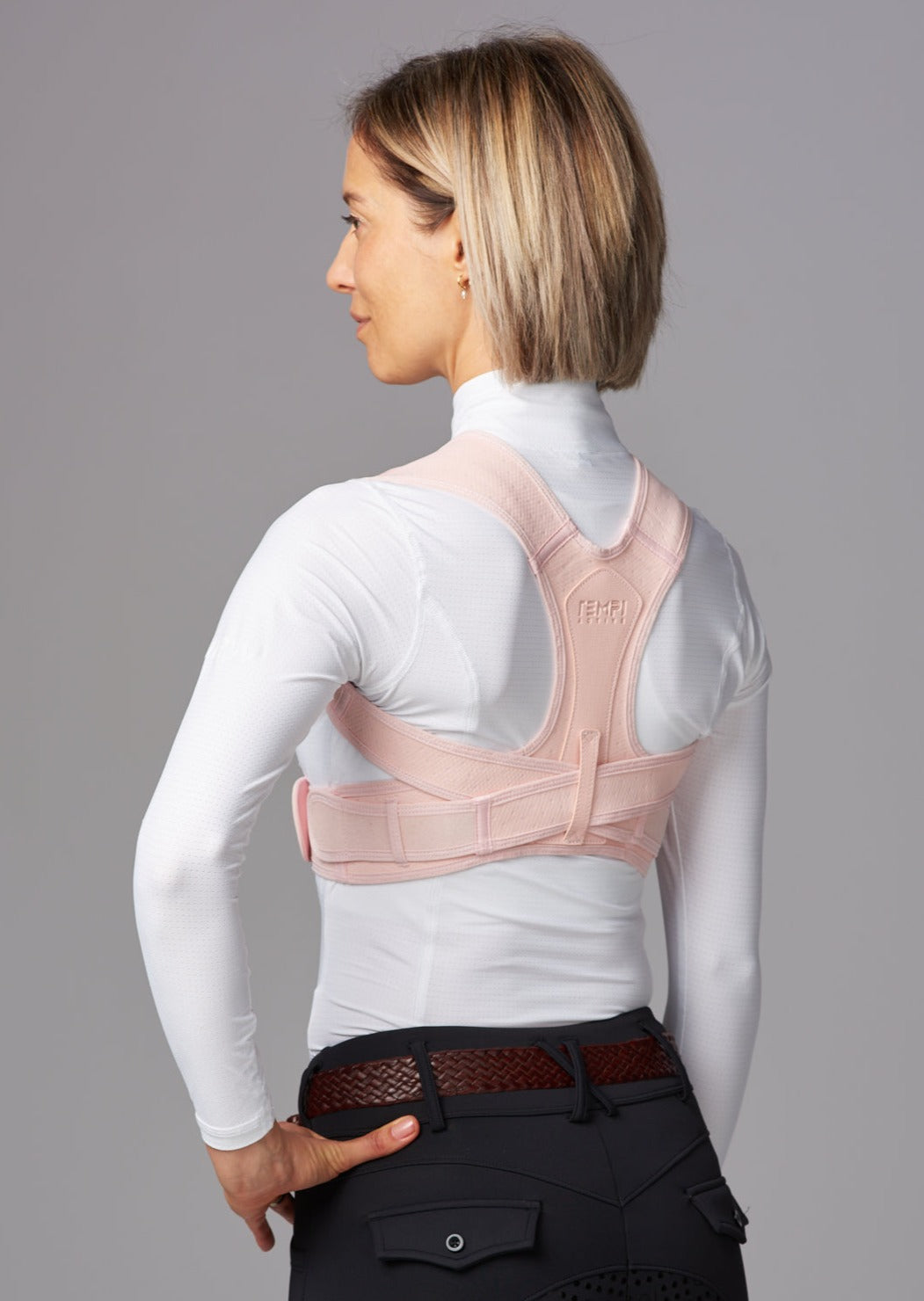 Female wearing dark riding pants, white top and brown leather belt, facing forward wearing pink posture brace.